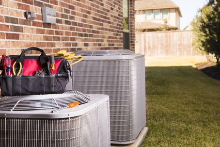 ag of repairman's work tools, gloves on top of two hvac units outside a brick home in a residential neighborhood.