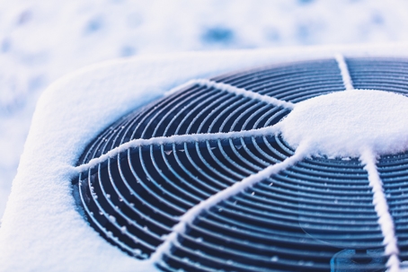 A close-up of an outdoor AC unit covered in snow, indicating freezing temperatures. | Blazer Heating, Air, and Plumbing